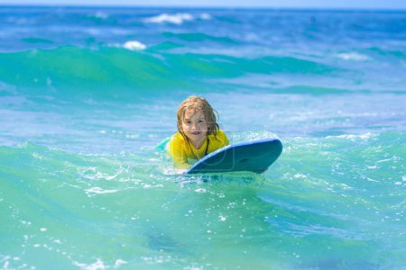 Foto de Los pequeños surfistas aprenden a montar en la tabla de surf en las olas del mar. Deportes acuáticos al aire libre para niños, actividad de natación en el campamento de surf. Vacaciones infantiles de verano en sat. Niño aprendiendo a surfear en el mar o en el océano - Imagen libre de derechos