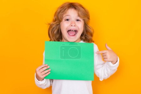 Photo for Amazed kid showing index finger on green empty sheet of paper, isolated on yellow background. Portrait of a kid holding a blank placard, poster. Surprised face, amazed emotions of child - Royalty Free Image