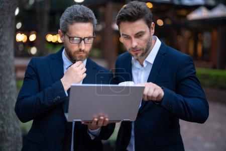 Photo for Two handsome young businessmen in classic suits using laptop. Businessmen discussing outdoor. Two business people talk project strategy. Two american businessmen in suits discuss business - Royalty Free Image