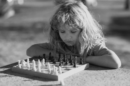 Téléchargez les photos : Pupil gamin penser à son prochain mouvement dans un jeu d'échecs. Petit garçon concentré assis à la table et jouant aux échecs - en image libre de droit