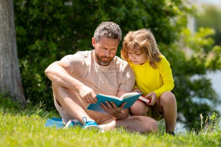 Photo for Father and son enjoy spending quality time reading books outdoors. Portrait of father and son relax on green lawn in summer park and read book together. Father and his little son reading book in park - Royalty Free Image