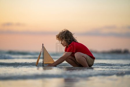 Foto de Niño con barco de juguete en agua de mar en las vacaciones de verano. Niño jugando con juguete barco de vela en el mar. Vacaciones de verano, niño jugando con el barco de juguete en la playa. Un chico con una nave de juguete. Día de los niños - Imagen libre de derechos