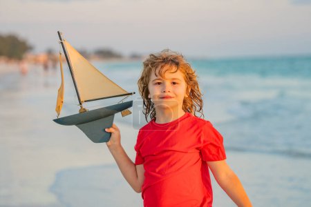 Téléchargez les photos : Enfant profitant de vacances d'été en bord de mer. Un petit garçon mignon jouant avec un bateau jouet sur la plage par une chaude journée ensoleillée d'été. Vacances en mer. Enfant heureux jouant avec un voilier jouet. Journée des enfants - en image libre de droit