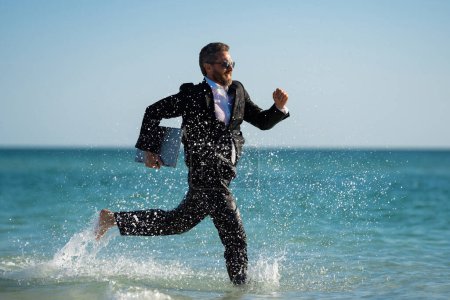 Photo for Business man in suit with laptop running in sea water. Travel tourism and business concept. Crazy male office employee with laptop running in sea. Crazy business man on summer vacation - Royalty Free Image
