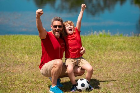 Photo for Dad with his little cute son are having fun and playing football on green grassy summer lawn. Football soccer sport concept. Father and son celebrate together after scoring goal or making a good play - Royalty Free Image