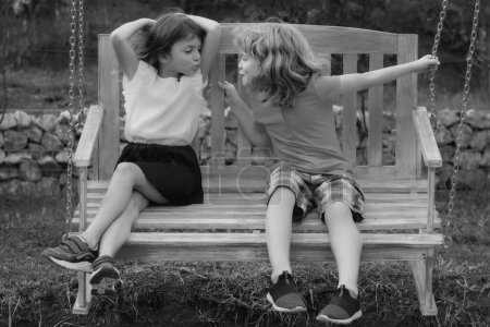 Foto de Dos niños al aire libre. Retrato de adorable hermano y hermana sonríen y ríen juntos mientras están sentados en columpio al aire libre. Niños felices balanceándose en jardín columpio de madera - Imagen libre de derechos