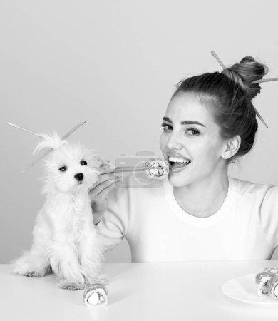 Foto de Hermosa chica comiendo rollos de sushi japonés con palillos, perro divertido, orden en el restaurante de comida rápida. Mujer de belleza con cachorro de mascota sosteniendo sushi con palillos - Imagen libre de derechos