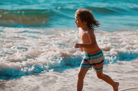 Téléchargez les photos : Enfant traversant l'eau près du rivage le long de la plage de la mer. Un garçon court le long de la côte. Reste des enfants en vacances d'été - en image libre de droit