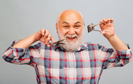 Téléchargez les photos : Vieux coiffeur. Heureux vieil homme barbu, homme barbu avec ciseaux et tondeuse à cheveux rasoir droite - en image libre de droit