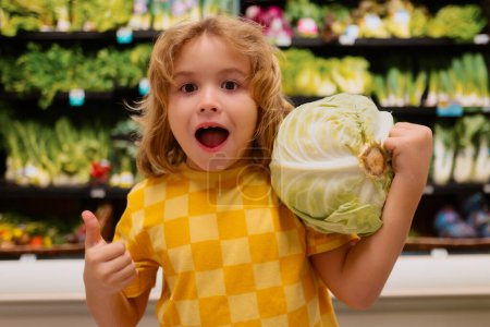 Foto de Niño con repollo. Compras en el supermercado. Niños comprando comestibles en el supermercado. El niño compra verduras frescas en la tienda de comestibles. Niño en la tienda - Imagen libre de derechos