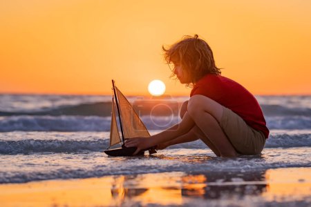 Téléchargez les photos : Petit garçon jouer avec le bateau jouet dans les vagues de la mer à la plage pendant les vacances d'été. Vacances d'enfance et d'été en famille. Rêve en voyage. Visite guidée en voilier. Concept de rêve enfants - en image libre de droit