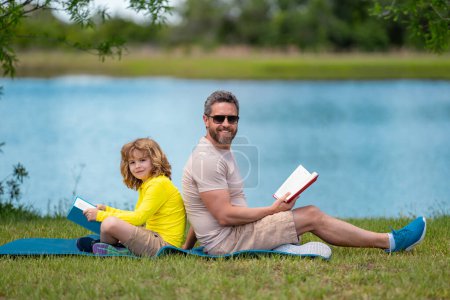 Téléchargez les photos : Père a lu un livre avec son fils dans un parc à l'extérieur. Père et fils enfant lisant en plein air sur fond de nature verte. Papa avec des enfants lisant un livre ensemble dans le parc d'été. Famille lisant un livre dans la nature - en image libre de droit