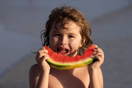 Téléchargez les photos : Il mange de la pastèque à la plage. Enfant tenant tranche de pastèque sur la plage. Vacances d'été amusant et concept de voyage. Enfant en mer pendant les vacances en plein air. Le gamin sourit, il aime manger des fruits - en image libre de droit