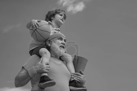 Photo for Preparing for morning workout in the park. Happy smiling boy on shoulder grandpa looking at camera. Child sits on the shoulders of his grandfather - Royalty Free Image