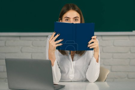 Photo for Student preparing exam and learning lessons in school classroom. Female college student hiding behind an open book and looking away. Girl covering face with book - Royalty Free Image