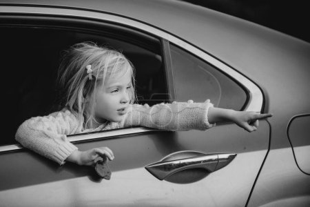 Téléchargez les photos : Petite fille dans la voiture montre quelque chose. Petite fille assise dans la voiture. Adorable petite fille regardant par la fenêtre de la voiture - en image libre de droit