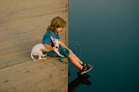 Foto de Divertido niño jugando con su perro cachorro en el parque cerca del lago. Una infancia sin preocupaciones. Protección animal doméstico - Imagen libre de derechos