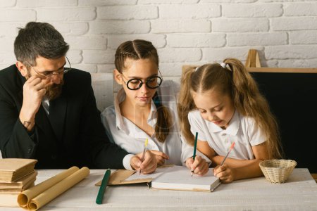 Photo for Elementary students with teacher in classroom. Father teaching schoolchild girls. Father and little kids daughters at school. Reading book and writing - Royalty Free Image