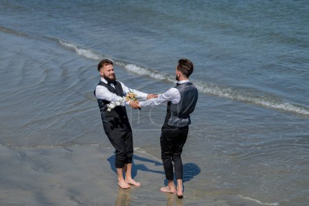 Foto de Feliz pareja gay masculina con flores cogidas de la mano en la boda. Pareja gay romántica y sensual. Los novios gay caminando juntos en la playa del mar durante el día de la boda. Hombres románticos en el agua de mar - Imagen libre de derechos