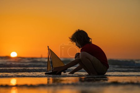Photo for Kid playing with toy sailing boat, toy ship. Travel and adventure concept. Child feeling adventurous while cruising. Kid on summer sunset beach. Kid playing on the beach at the sunset time - Royalty Free Image