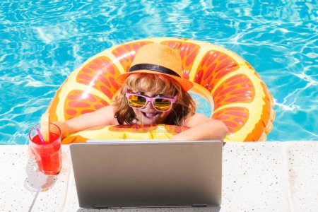 Téléchargez les photos : Boire un cocktail d'été pour enfants et utiliser un ordinateur portable dans la piscine. Travail indépendant, enseignement en ligne à distance, apprentissage en ligne. Cours d'été. Enfant apprentissage à distance sur ordinateur portable dans la piscine - en image libre de droit