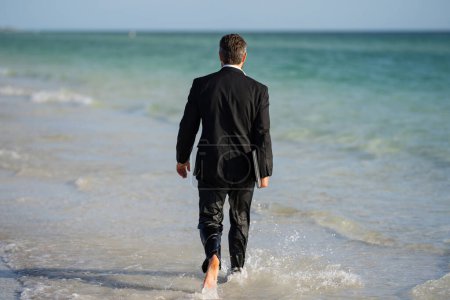 Téléchargez les photos : Vue arrière du dos homme d'affaires en costume dans l'eau de mer à la plage. Beau homme d'affaires en vacances d'été. Homme d'affaires en cours d'exécution sur la mer tenant ordinateur portable. Homme d'affaires sautant dans la mer - en image libre de droit