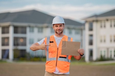 Photo for Engineer hold paper board for text. Builder showing signboard placard banner, poster. Serious engineer at new home. Construction manager in helmet - Royalty Free Image