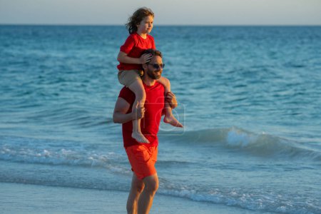 Photo for Outdoor portrait of happy father and son walking on sunny ocean beach. Father and son walk in sea beach. Son on fathers shoulders piggyback ride. Family on beach, summer holiday vacation - Royalty Free Image