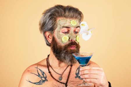 Téléchargez les photos : Spa homme avec masque d'argile et tranches de concombre sur cocktail boisson visage. Spa, dermatologie, concept de bien-être et de soins du visage. Homme ayant masque hydratant cosmétique, fond studio isolé - en image libre de droit