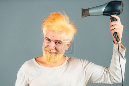Foto de Hombre pelirrojo guapo con el pelo largo seca su cabello con un secador de pelo. Hombre rubio con secador de pelo - Imagen libre de derechos