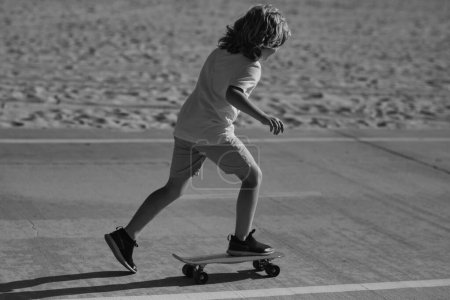 Téléchargez les photos : Kid skateboarder monter un skateboard dans la rue. Enfant dans une ville d'été. Petit enfant, tout-petit garçon à cheval skateboard dans le parc pour la première fois, essayer le skateboard - en image libre de droit
