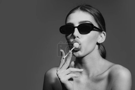 Photo for Lollypop and woman face, Sexy woman in sunglasses with a lollipop on studio background. Fashion model portrait licking candy - Royalty Free Image