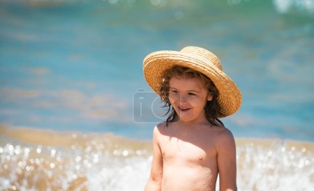 Foto de Adorable niño en sombrero de paja en la playa durante las vacaciones de verano. Retrato de un niño juguetón sobre el fondo del mar. Divertido verano cara de niño - Imagen libre de derechos