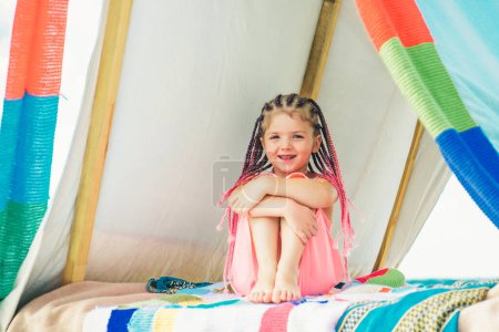 Téléchargez les photos : Un enfant heureux avec des dreadlocks joue dans une tente. Concept de camping pour enfants. Une fille qui joue au camp. S'amuser à l'extérieur au camping - en image libre de droit