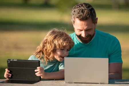 Téléchargez les photos : Père heureux utilisant ordinateur portable se détendre avec le fils écolier tenant ordinateur portable s'amuser ensemble, souriant papa et petit garçon enfant profiter week-end avec des gadgets à l'extérieur sur la nature - en image libre de droit
