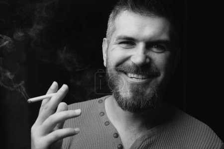 Téléchargez les photos : Un homme souriant fumant une cigarette. Fumée sur fond noir. Dépendance au tabac. Dépendance à la nicotine. Fumée sur noir - en image libre de droit