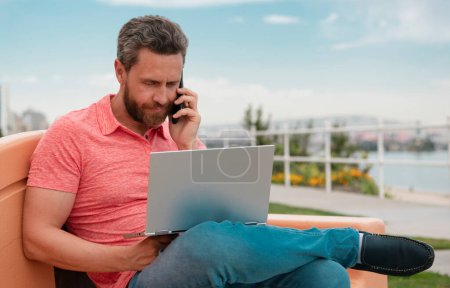 Téléchargez les photos : Beau jeune homme d'affaires travaillant avec ordinateur portable en plein air parler sur un téléphone mobile - en image libre de droit