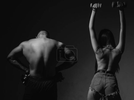 Foto de Muscular man with naked body, fitness woman with dumbbells on a dark background, back view. Sexy sport couple exercising with dumbbell. Slim and healthy sexy girl with strong muscular man workout - Imagen libre de derechos