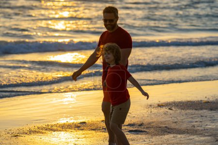 Téléchargez les photos : Père et fils se tenant la main marchant sur la plage du coucher du soleil. La fête des pères. Père marchant avec un petit enfant fils sur la plage près de la mer. Papa avec son fils marchant sur une plage d'été. Les pères aiment les enfants - en image libre de droit