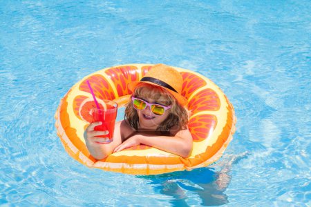 Foto de Niño en gafas de sol y sombrero de verano beber cóctel de verano y flotando en la piscina. Lindo niño pequeño relajándose con anillo de juguete flotando en una piscina divirtiéndose en las vacaciones de verano - Imagen libre de derechos