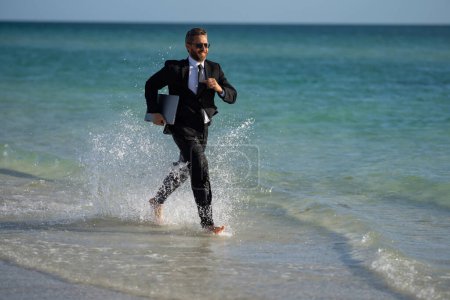 Foto de Turista hombre de negocios en traje casual descalzo caminando en la playa tropical con portátil. Hombre de negocios guapo en vacaciones de verano. Empresario corriendo en el mar sosteniendo el ordenador portátil. Empresario saltando en el mar - Imagen libre de derechos