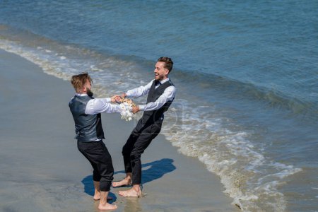 Foto de Retrato de feliz pareja gay enamorada el día de la boda. Los novios gay caminando juntos en la playa del mar durante el día de la boda. Hombres románticos en el agua de mar - Imagen libre de derechos