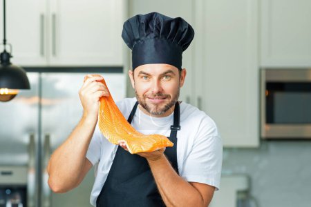 Foto de Hombre guapo cocinando salmón de pescado en la cocina. Retrato de un hombre casual cocinando en la cocina con salmón. Hombre casual preparando salmón de pescado crudo en la cocina. Chef con filete de salmón grande - Imagen libre de derechos