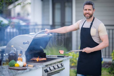 Téléchargez les photos : Beau mâle préparant barbecue, grill en plein air. Homme cuisiner de la viande et du poisson sur le barbecue dans la cour arrière. Cuisinière au barbecue. Barbecue de cuisson. Préparation de la nourriture sur le gril à l'aide d'un ensemble barbecue - en image libre de droit
