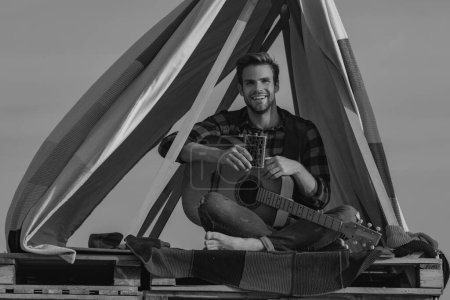 Photo for Music outdoors. Sexy man with guitar in checkered shirt, hipster camping and hiking. Man with bare muscular torsowith guitar. Man playing guitar and sitting near the tent in the camp - Royalty Free Image
