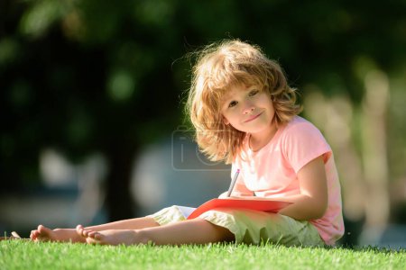 Téléchargez les photos : Joyeux élève préscolaire dans le parc sur l'herbe. Éducation des enfants en plein air. Concept d'éducation et d'apprentissage des enfants - en image libre de droit