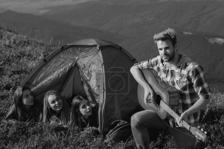 Téléchargez les photos : Groupe de jeunes femmes gaies dans la tente du camp ensemble tandis que jeune bel homme jouant de la guitare - en image libre de droit