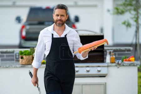 Foto de Chef masculino en delantal de cocinero asando y parrillando salmón pescado en el jardín. Hombre guapo preparando filete de salmón asado. Barbacoa y parrilla. Cocinar pescado en el patio trasero - Imagen libre de derechos