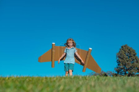 Téléchargez les photos : Child boy plays in an astronaut dreams of space. Happy child play with toy plane cardboard wings against blue sky. Kid having fun in summer field outdoor. Portrait of boy with paper wings - en image libre de droit