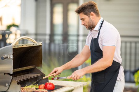 Téléchargez les photos : Beau homme préparant barbecue grill extérieur. Homme cuisiner des aliments savoureux sur barbecue grill à l'arrière-cour. Chef préparant la nourriture sur barbecue. Homme millénaire griller de la viande sur le gril. Bbq party. Grillades de repas - en image libre de droit